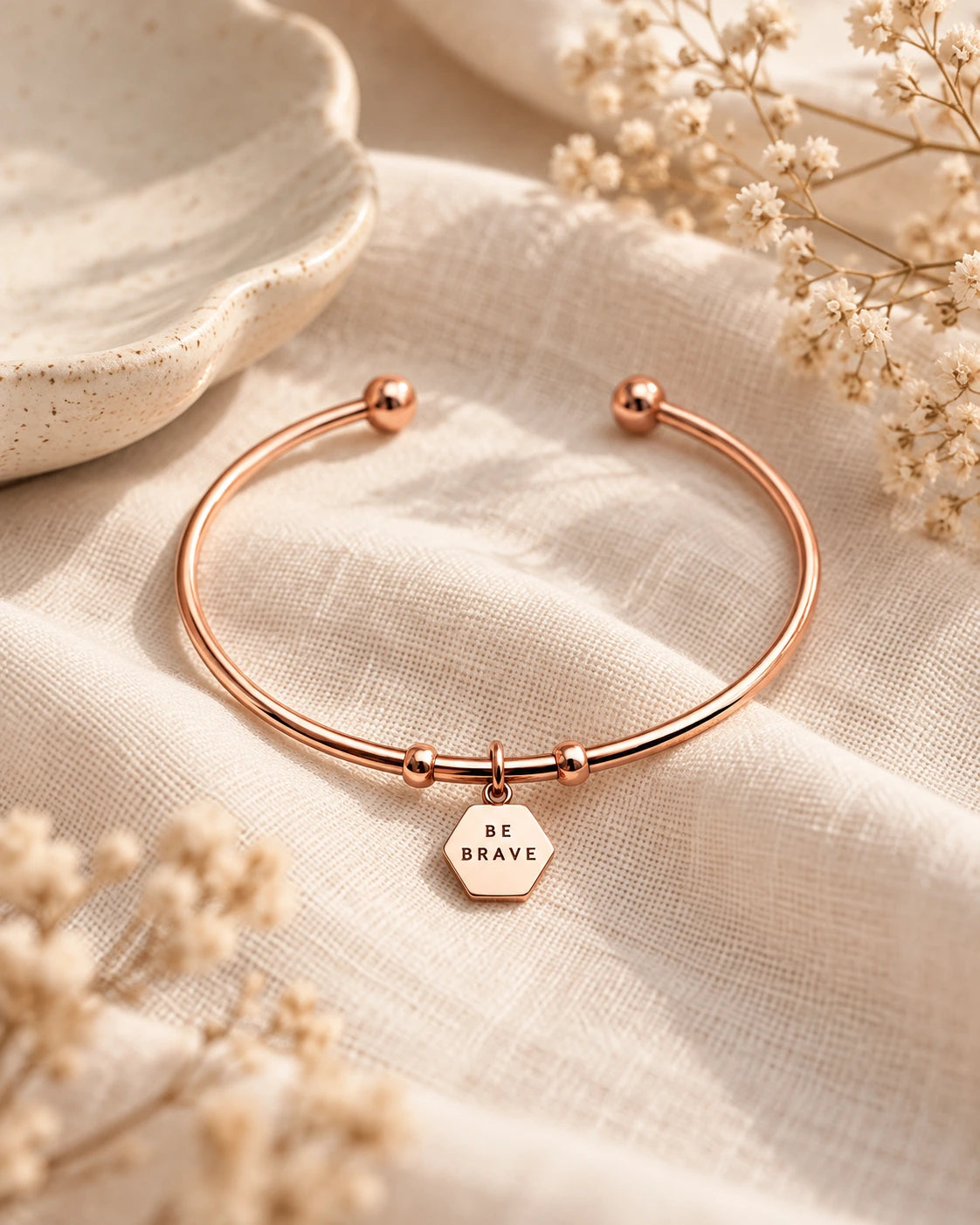 Rose gold hexagon pendant bracelet engraved Be Brave styled on soft cream linen with dried flowers