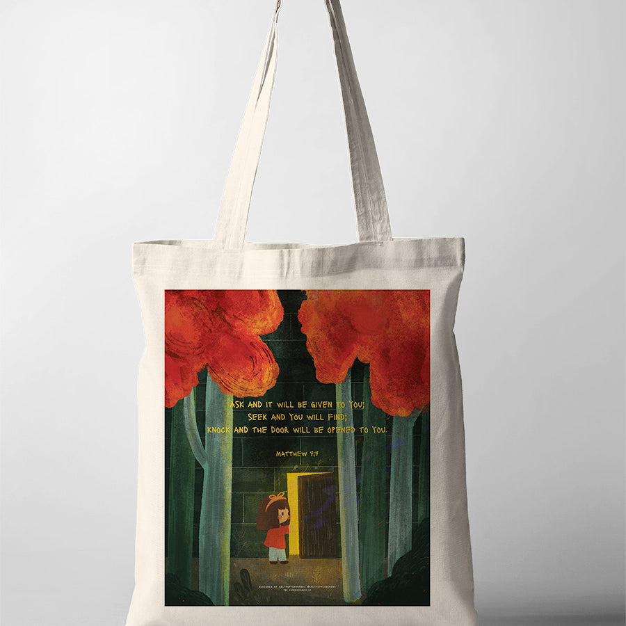 A beige tote bag featuring a Christian-themed illustration of a child standing in front of an open door in a forest, with red trees above. The design includes the Bible verse from Matthew 7:7: 'Ask and it will be given to you; seek and you will find; knock and the door will be opened to you,' printed on the bag, symbolizing faith and guidance.