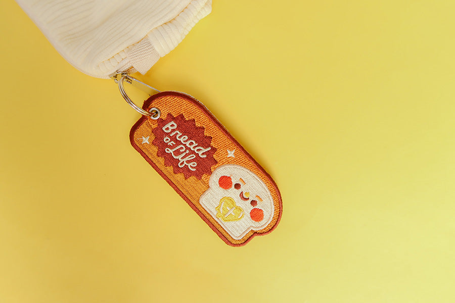 Brown embroidered keychain with a cheerful bread character and the text 'Bread of Life'.