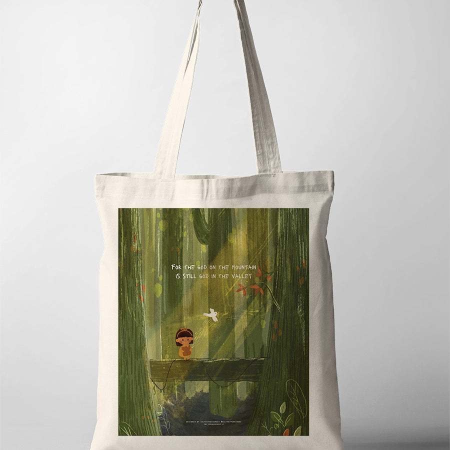 A beige tote bag featuring a Christian-themed illustration of a child standing on a bridge in a forest, with a white bird flying nearby. The design includes the text, 'For the God on the mountain is still God in the valley,' conveying a message of faith and God's presence in all circumstances.