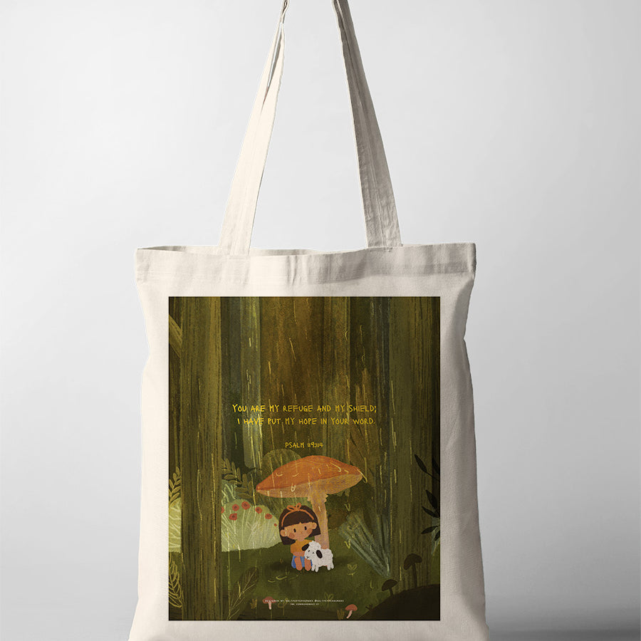 A beige tote bag featuring a Christian-themed illustration of a child sitting under a large mushroom with a small white dog. The design includes the Bible verse from Psalm 119:114: 'You are my refuge and my shield; I have put my hope in your word,' conveying a message of comfort and faith.