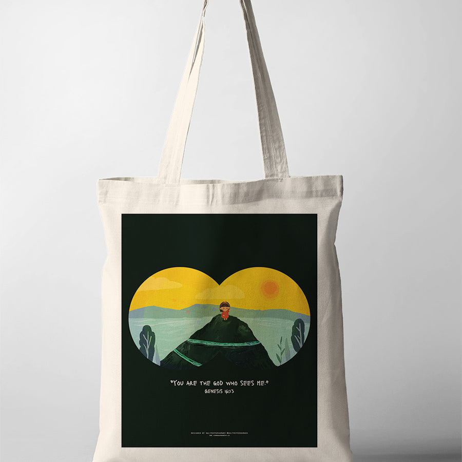 A beige tote bag featuring an illustration of a child sitting on a hilltop with a vibrant yellow and green landscape in the background, symbolized through binoculars. The design includes the Bible verse from Genesis 16:13: 'You are the God who sees me,' conveying a message of being seen and known by God.