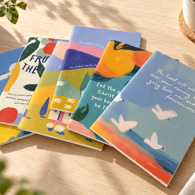 Lifestyle photo of colourful A6 Christian notebooks arranged on a sunlit wooden desk with plants and a coffee cup