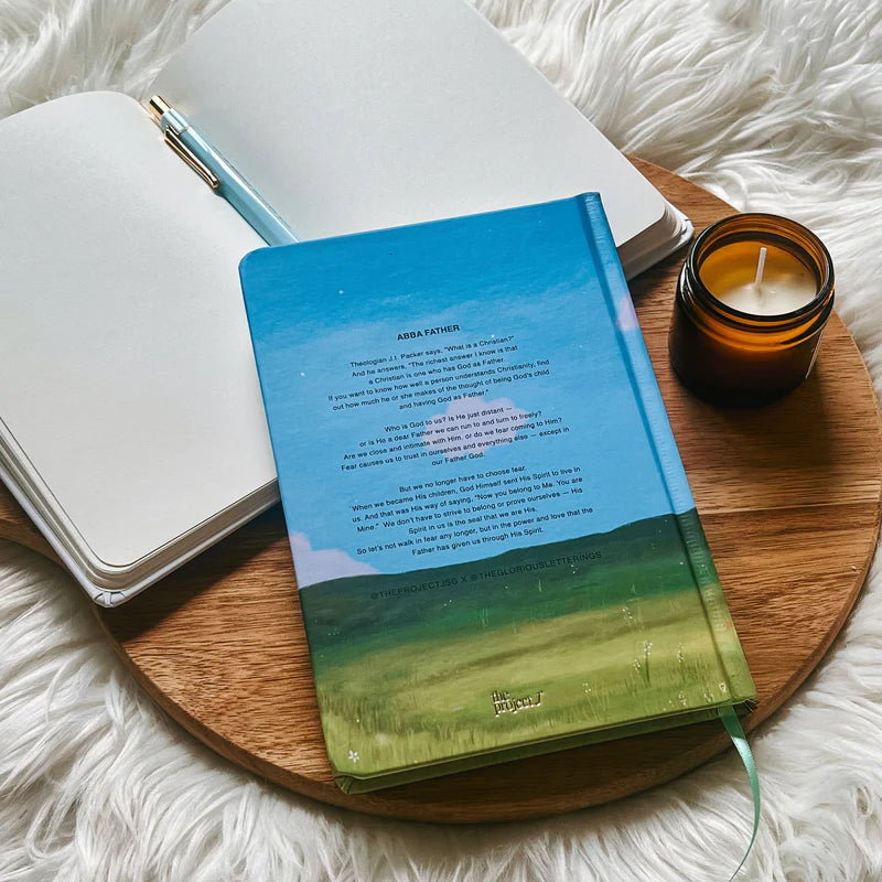 Hardcover journal with printed message on back cover by Project J placed on wooden tray beside open notebook and candle.