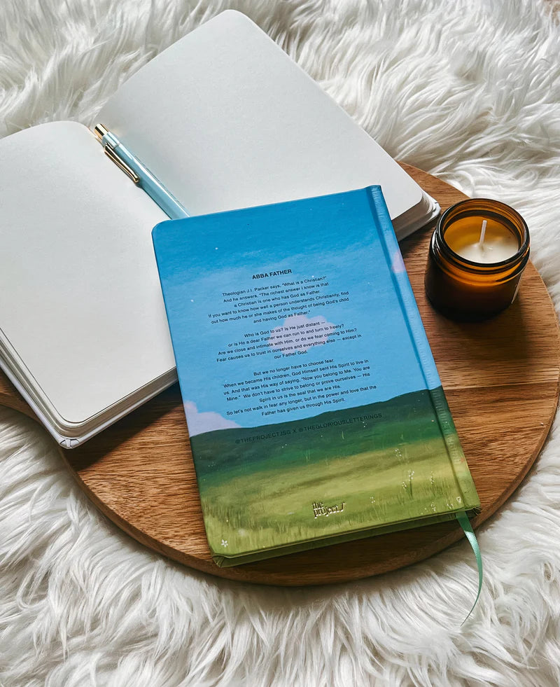 Hardcover journal with printed message on back cover by Project J placed on wooden tray beside open notebook and candle.