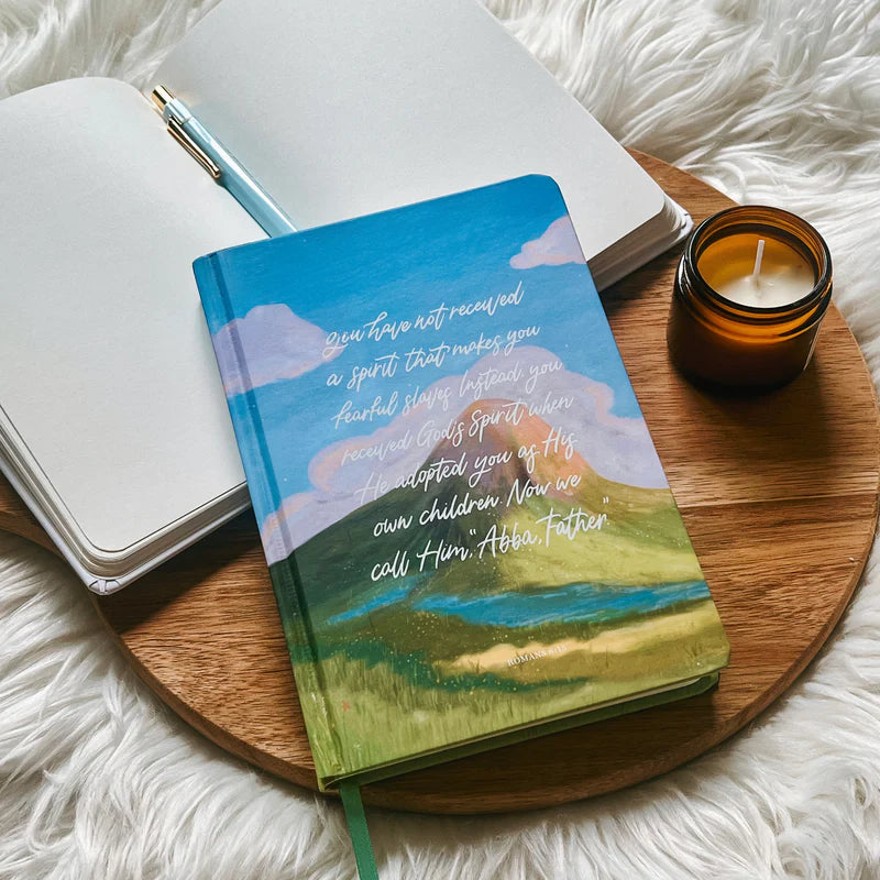 Hardcover journal with Romans 8 verse and painted sky design by Project J placed on wooden tray beside open notebook and candle.