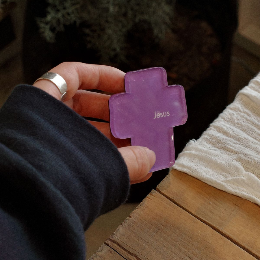 A hand holding a vibrant purple acrylic magnet shaped like a cross with the name "Jesus" inscribed on it, set against a softly blurred background of greenery.