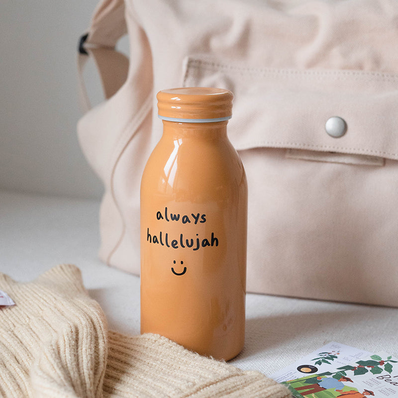 Mustard-colored milk bottle tumbler with the handwritten phrase "always hallelujah" and a smiley face, set on a light surface with a beige bag and cozy sweater in the background.