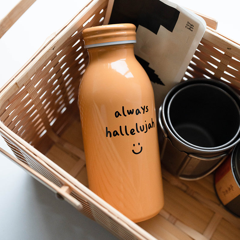 Mustard-colored milk bottle tumbler with the handwritten phrase "always hallelujah" and a smiley face, shown in a wooden gift basket.