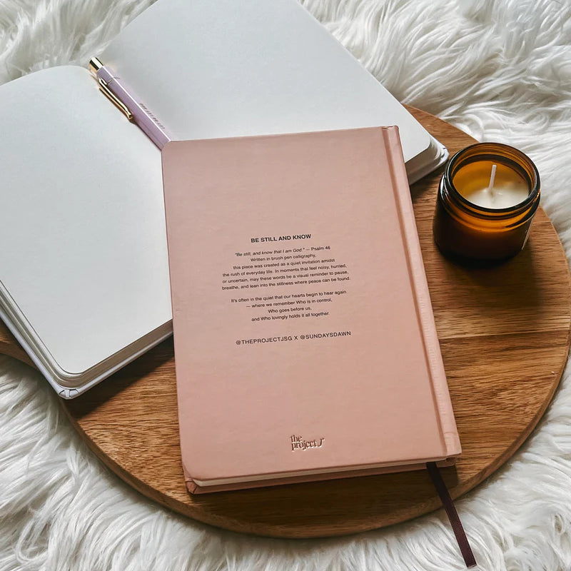 Blush pink hardcover journal with printed message on back cover by Project J placed on wooden tray beside open notebook and candle.
