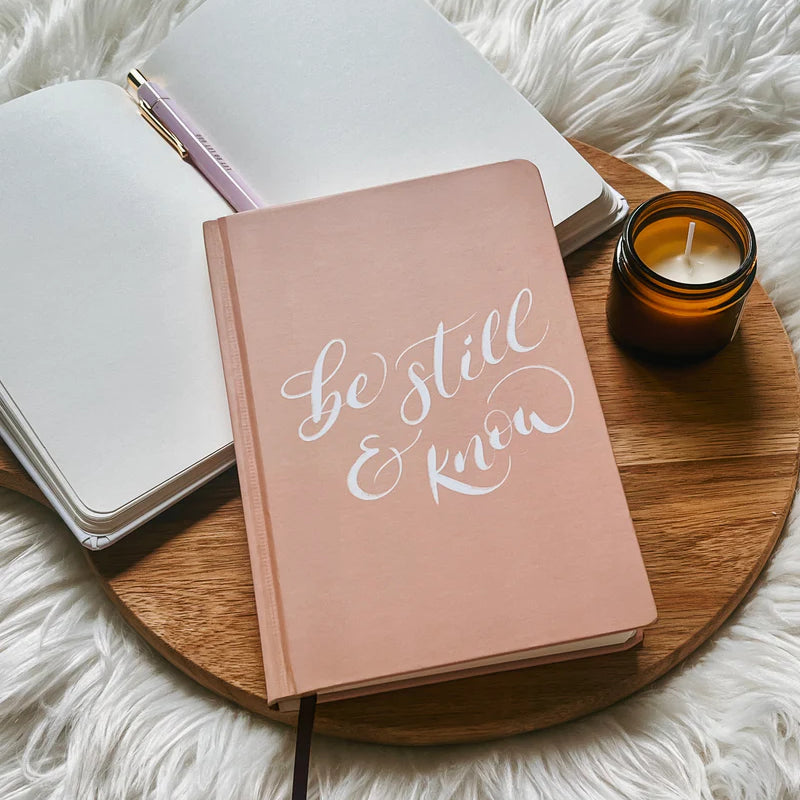 Blush pink hardcover journal with “Be still & know” text by Project J placed on wooden tray beside open notebook and candle.