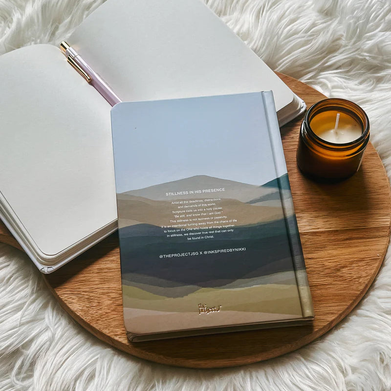 Hardcover journal with printed reflection text on back cover by Project J placed on wooden tray beside open notebook and candle.