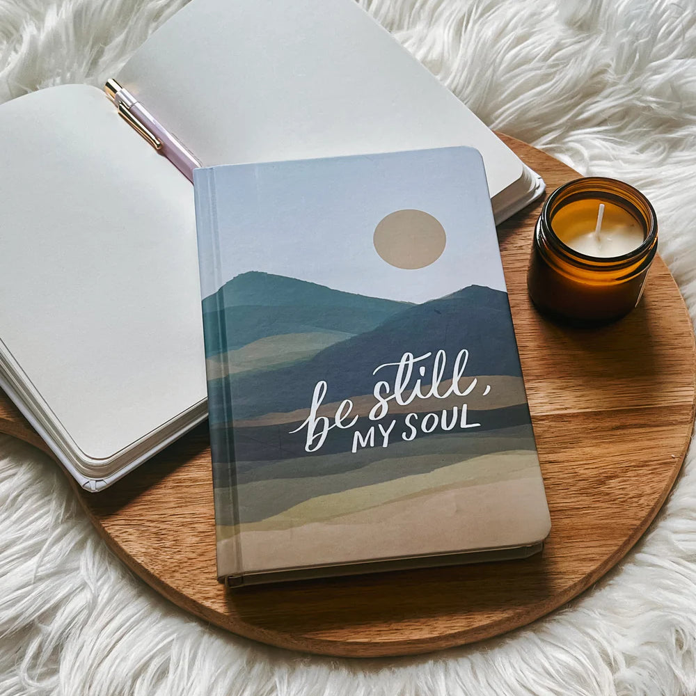 Hardcover journal with “Be still, my soul” lettering design by Project J placed on wooden tray beside open notebook and candle.