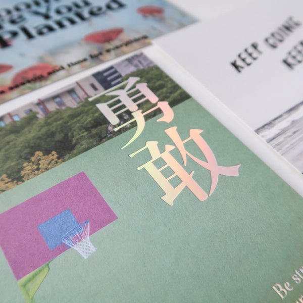 A close-up of a collection of postcards with various inspirational messages, including a card with the Chinese characters for "courage" (勇敢) and the text "Be strong & courageous," alongside other motivational cards like "Keep Going, Keep Loving" and "Bloom Where You Are Planted."