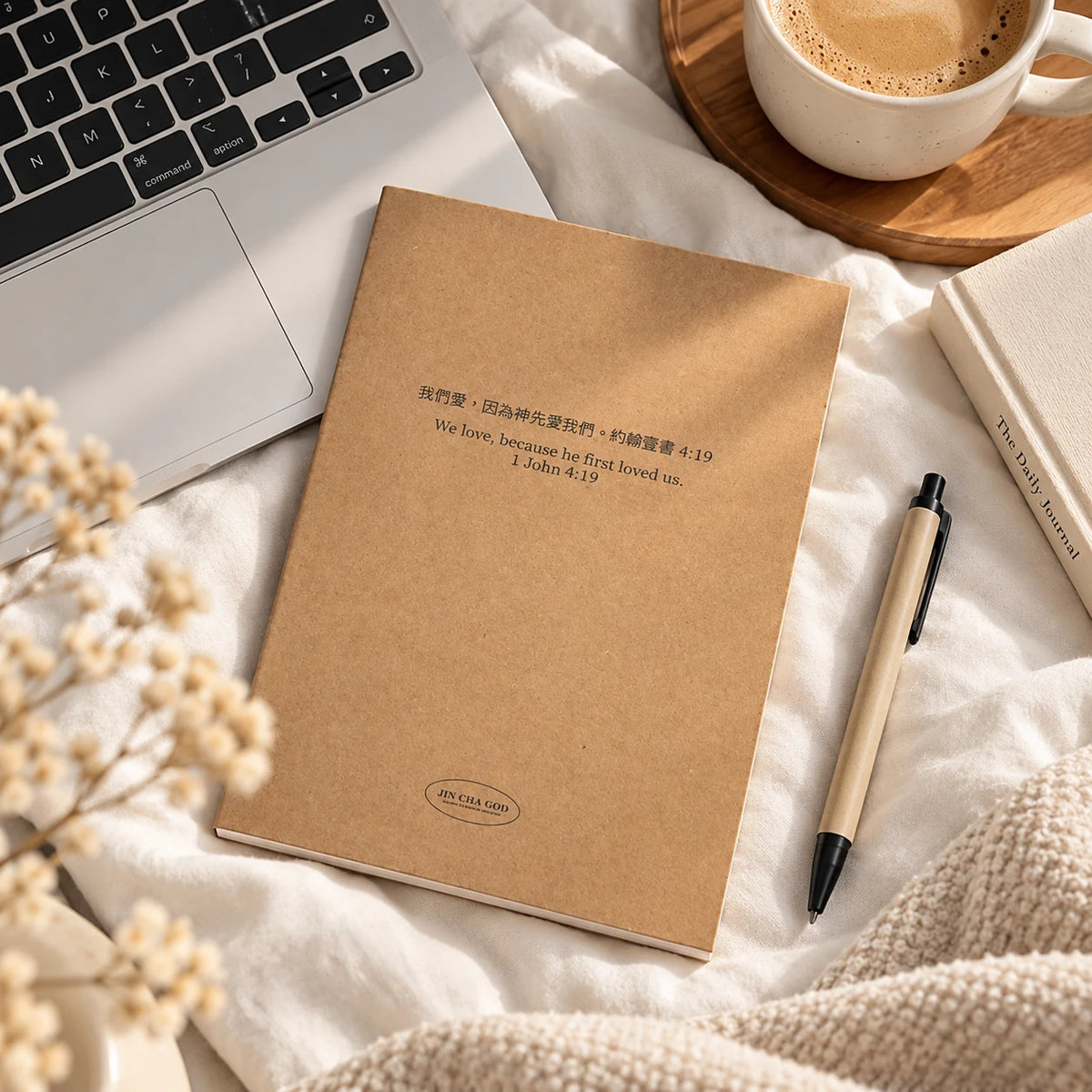 Because He First Loved Us 1 John 4:19 A5 kraft notebook lying on cream bedding beside a laptop and coffee