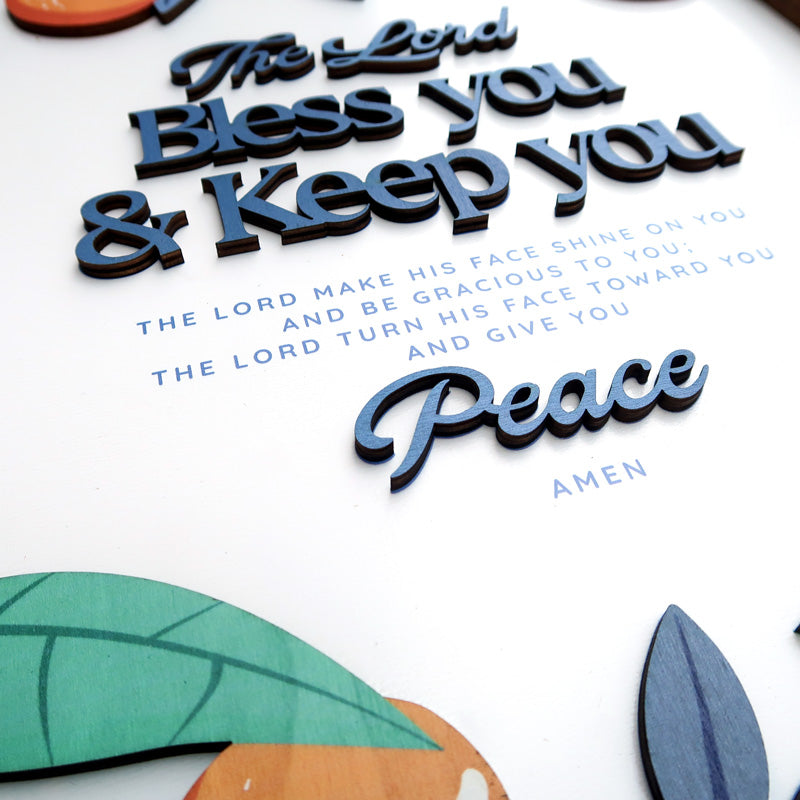 Close-up of a handcrafted wooden scripture poster featuring the phrase "The Lord Bless You and Keep You" with "Peace" in a layered design. The poster includes colorful wooden elements and a natural theme, framed in wood.