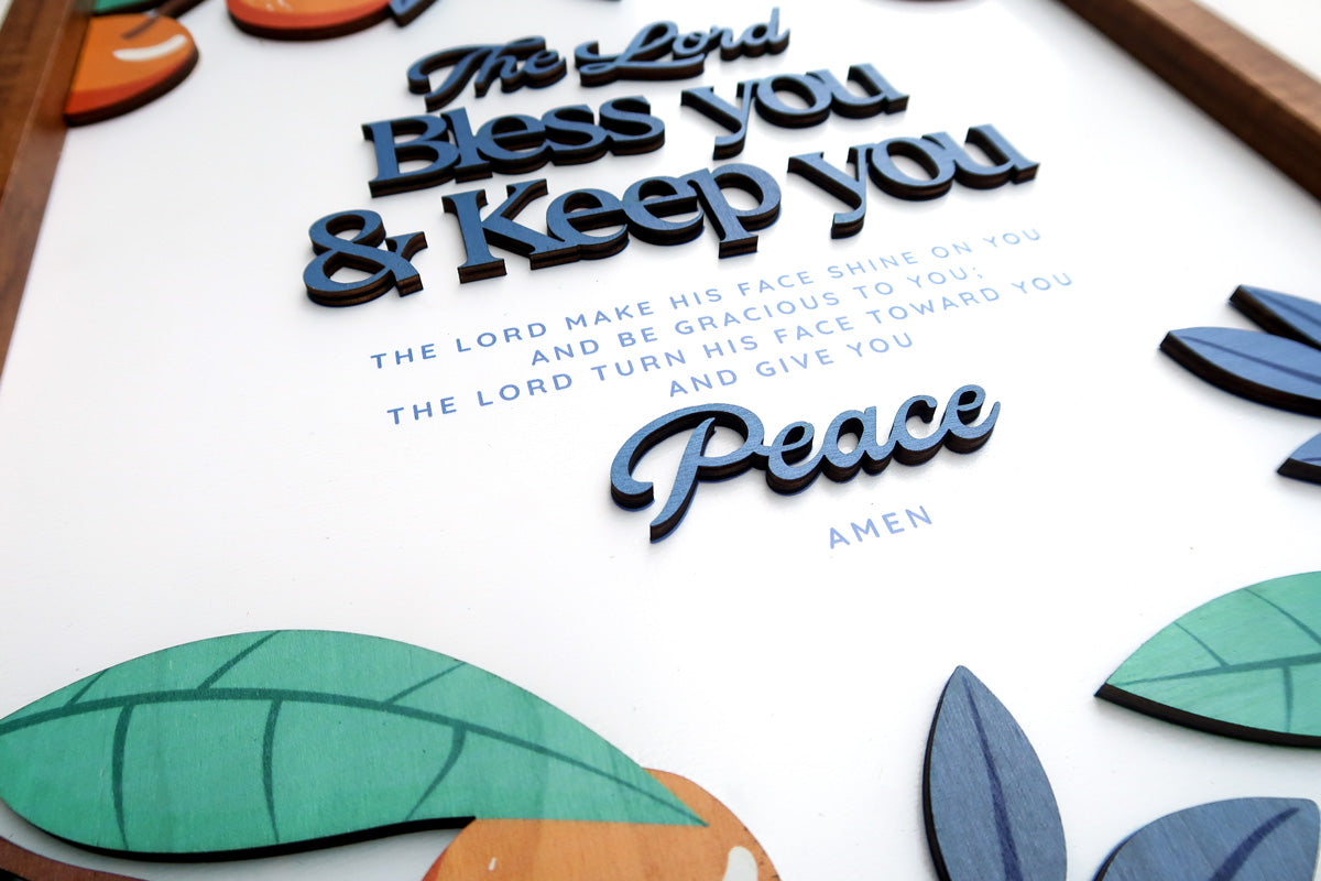 Close-up of a handcrafted wooden scripture poster featuring the phrase "The Lord Bless You and Keep You" with "Peace" in a layered design. The poster includes colorful wooden elements and a natural theme, framed in wood.