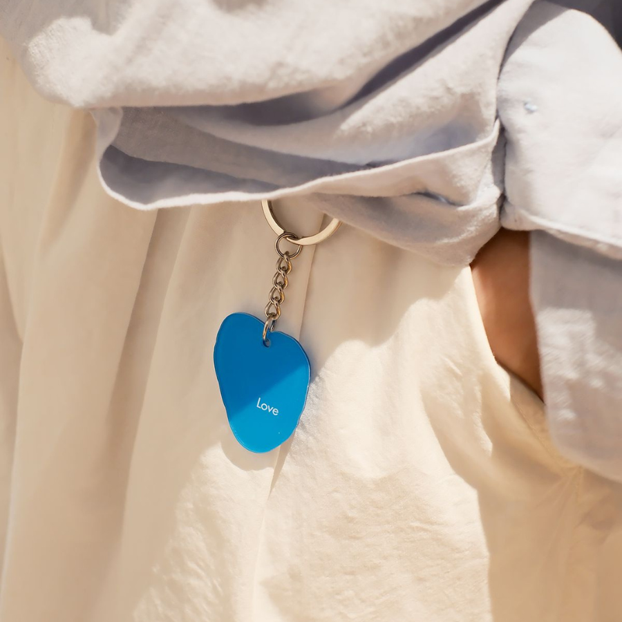 Blue heart-shaped keychain with "Love" inscribed, attached to a light-colored fabric, symbolizing faith and affection.