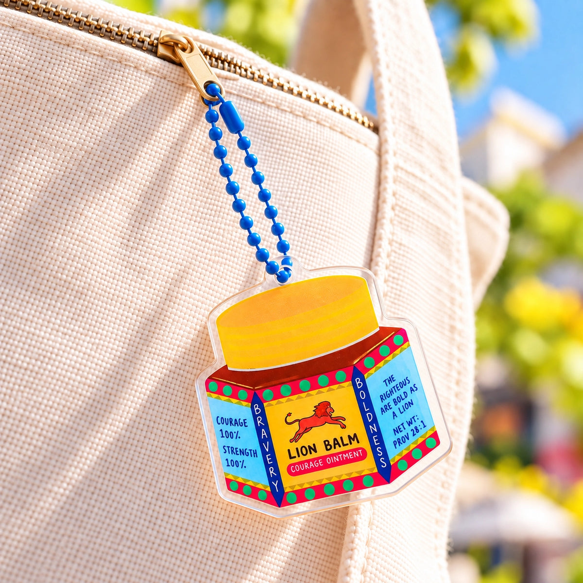 Bold Lion Balm Christian acrylic keychain attached to a cream tote bag in a sunny outdoor setting.