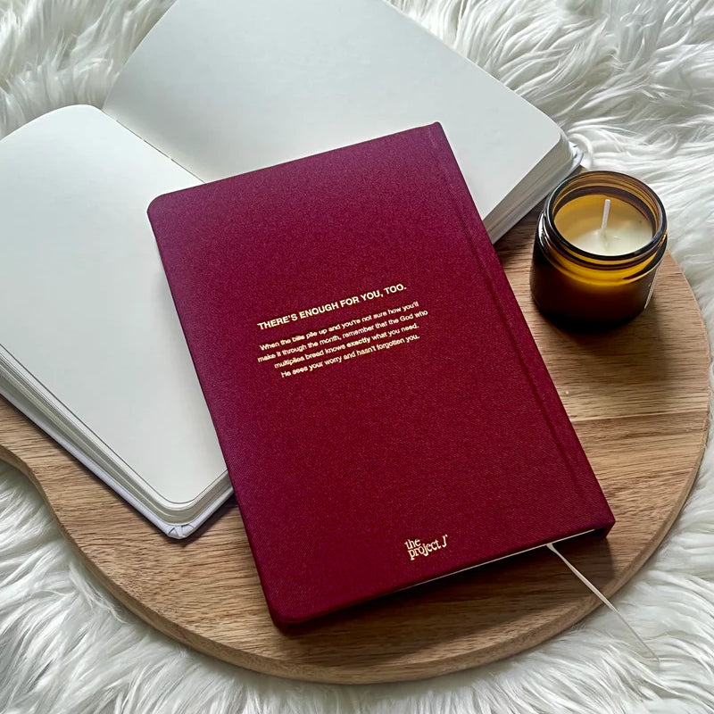 Red fabric journal with gold quote on back cover by Project J placed on wooden tray beside open notebook and candle.