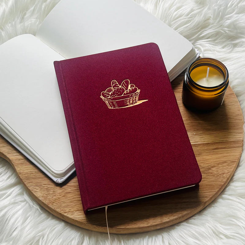 Red fabric journal with gold bread basket design by Project J placed on wooden tray beside open notebook and candle.