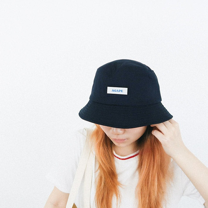 A person wearing a navy blue bucket hat featuring a white patch with the word "AGAPE" in blue, showcasing a faith-inspired and customizable streetwear accessory.