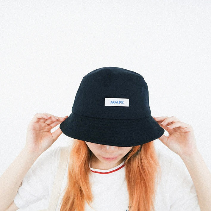 A person wearing a navy black bucket hat featuring a white patch with the word "AGAPE" in blue, showcasing a faith-inspired, modern streetwear accessory.