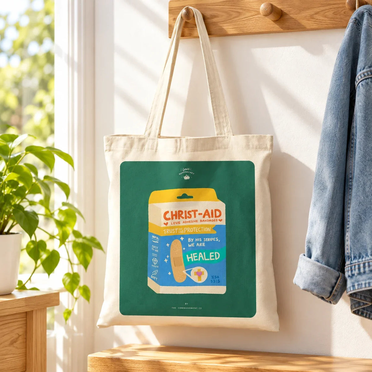 Christ-aid Love Bandages canvas tote bag on a wooden peg in a sunlit home entryway.