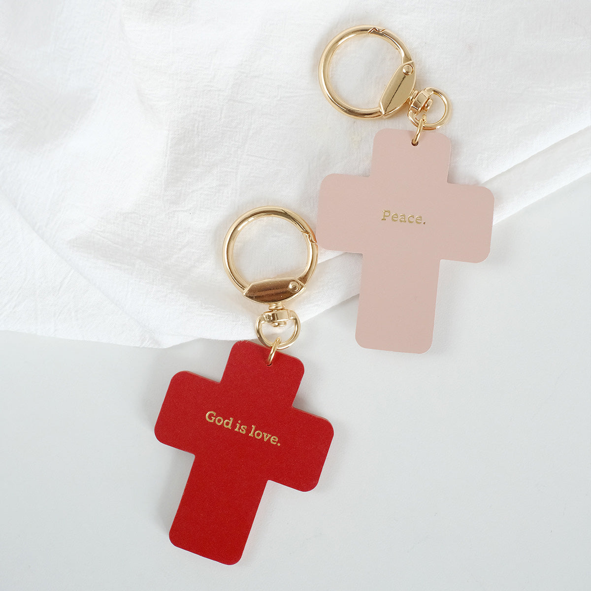 Two cross-shaped keychains with gold rings, one red with 'God is love' and one pink with 'Peace,' displayed on a white background.