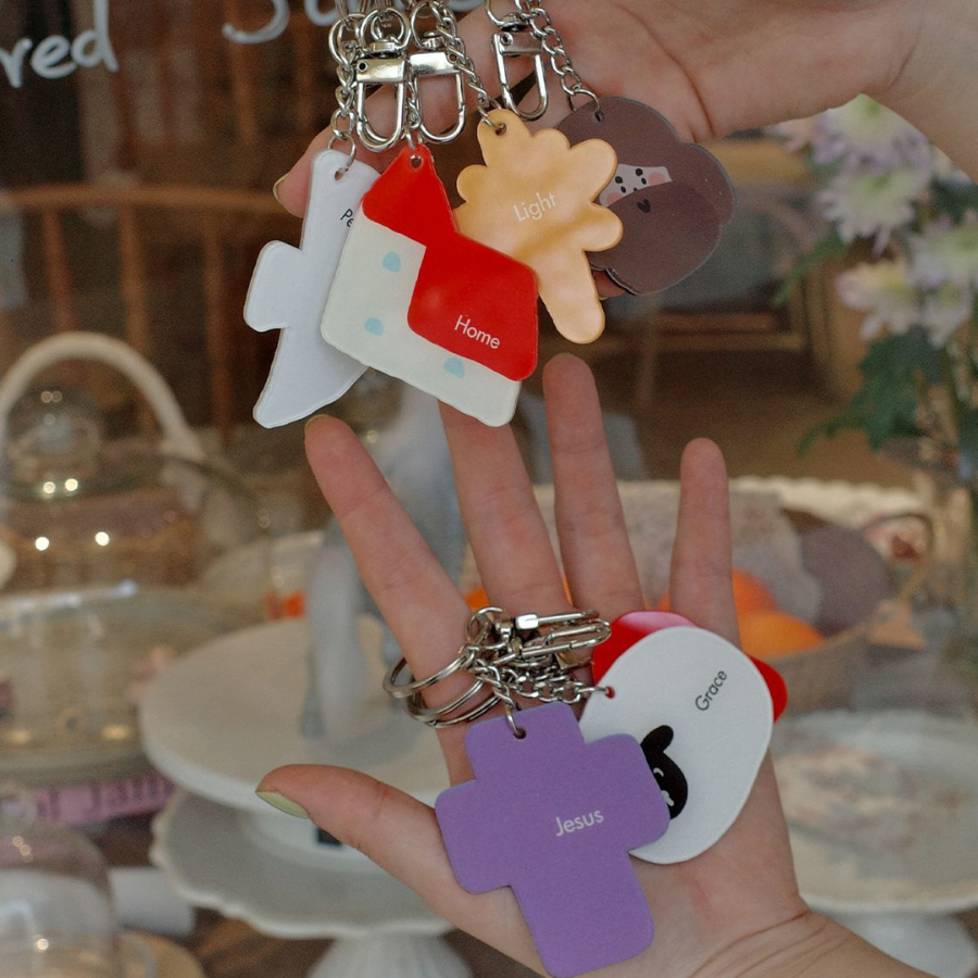 A variety of Christian-themed keychains with words like "Jesus," "Home," and "Light," held in a hand, showcasing faith-inspired designs.