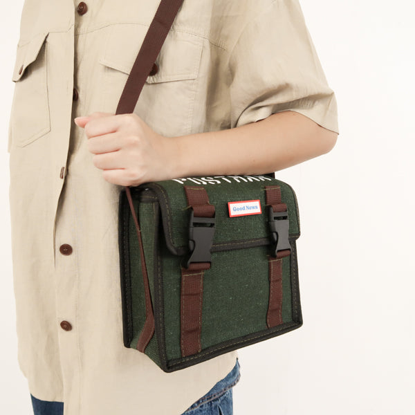 A close-up view of a person wearing a green canvas messenger bag with 'Good News' branding, highlighting the crossbody strap and faith-based design. The bag features sturdy brown straps and a durable canvas construction.