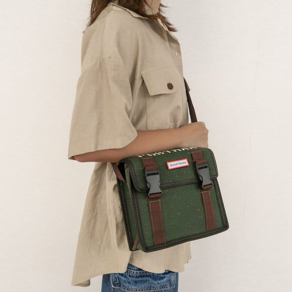 A person wearing a green canvas messenger bag with 'Good News' branding, shown from the side. The bag features brown straps and buckles, with a practical and faith-inspired design.