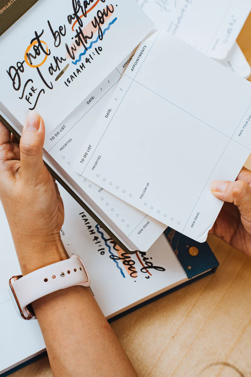 Hands holding a page from The Project J Christian notepad featuring the encouraging verse "Do not be afraid, for I am with you" from Isaiah 41:10, surrounded by faith-inspired planners and notes for uplifting daily organization.
