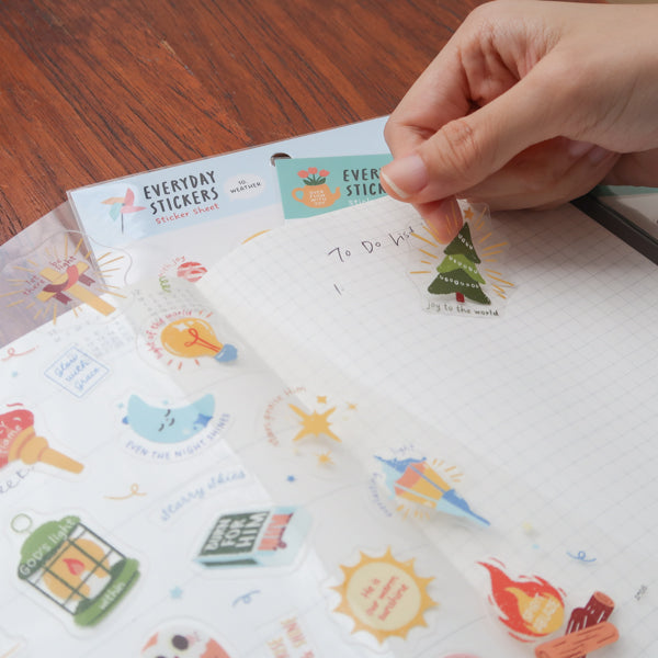 Hands peeling a Christian sticker from everyday believer sticker sheet featuring light and faith-themed illustrations like lamp and stars
