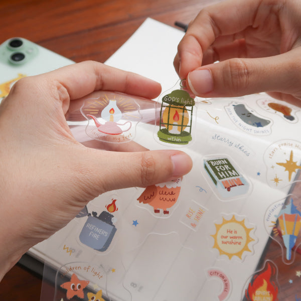 Hands peeling a Christian sticker from everyday believer sticker sheet featuring light and faith-themed illustrations like lamp and stars
