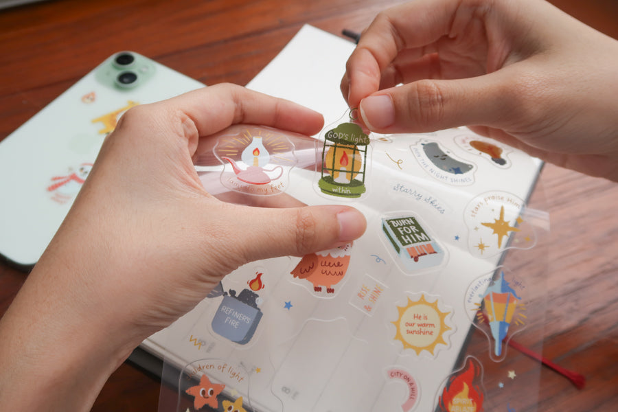 Hands peeling a Christian sticker from everyday believer sticker sheet featuring light and faith-themed illustrations like lamp and stars