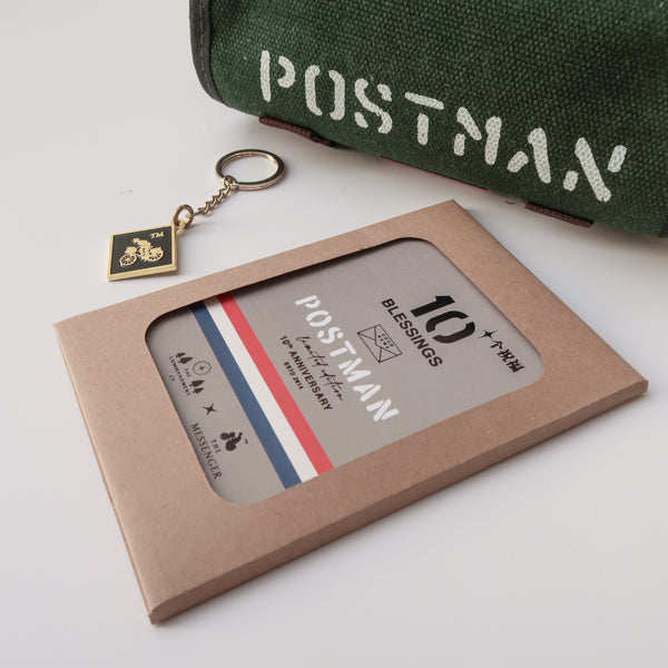 A close-up of the 'Good News' green canvas messenger bag with 'POSTMAN' text, displayed with a 10th anniversary edition 'Postman' keychain and blessings packaging. The keychain features a motorbike design, complementing the faith-inspired product set.