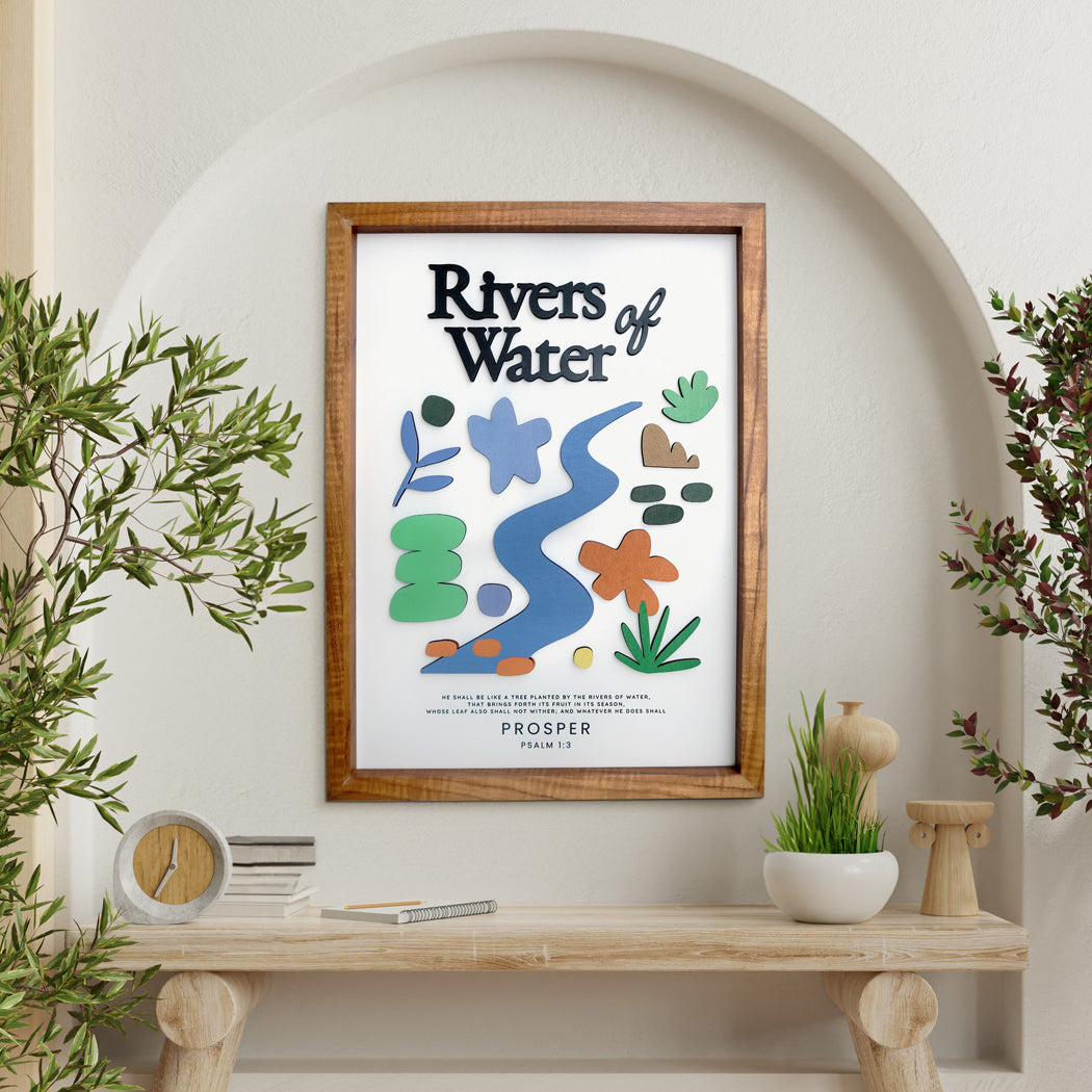 A modern wooden-framed poster featuring the Bible verse "Rivers of Water - Psalm 1:3" with colorful abstract and floral cutouts. The artwork is displayed in a bright, arch-shaped alcove surrounded by natural elements like potted plants and a wooden console table, creating a peaceful and elegant setting.