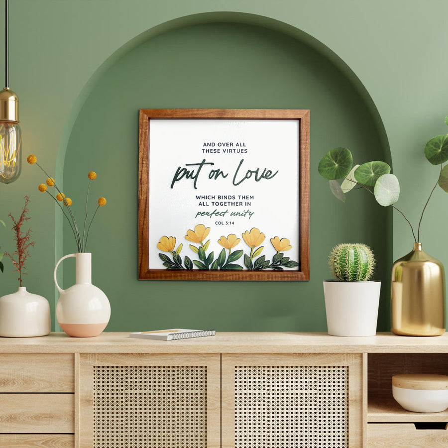 A beautifully crafted wooden-framed wall art featuring the Bible verse "Put on Love - Colossians 3:14." The poster includes yellow floral designs and an elegant script style. Displayed in a modern setting with green walls, an arched alcove, and decor elements like a wooden console, potted plants, and warm lighting, creating a serene and sophisticated ambiance.