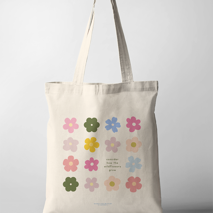 Neutral canvas tote bag decorated with colorful wildflower illustrations and the uplifting phrase "consider how the wildflowers grow," embodying simple happiness and faith-inspired encouragement.
