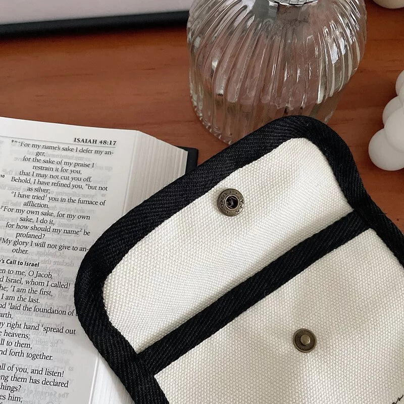 Cream canvas wallet with black trim and snap closure on a wooden desk beside an open Bible