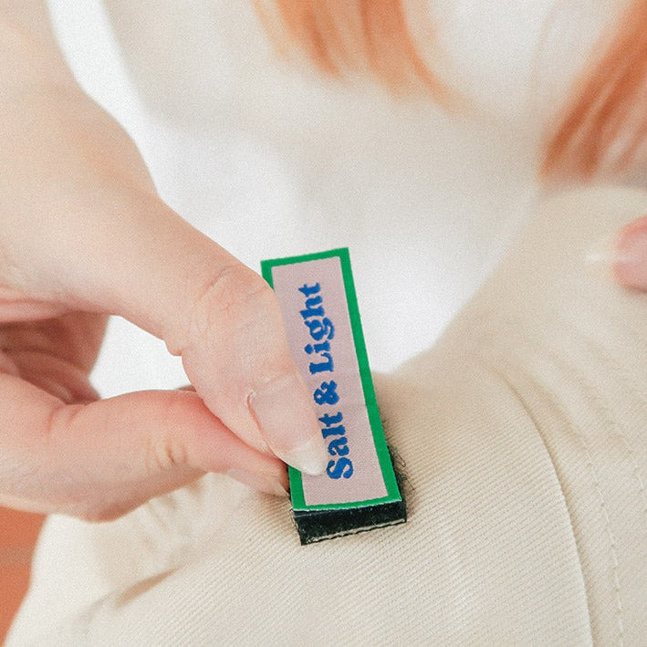 A close-up of a hand attaching a green and white velcro patch labeled "Salt & Light" onto a beige cap, showcasing customizable and interchangeable faith-based designs.