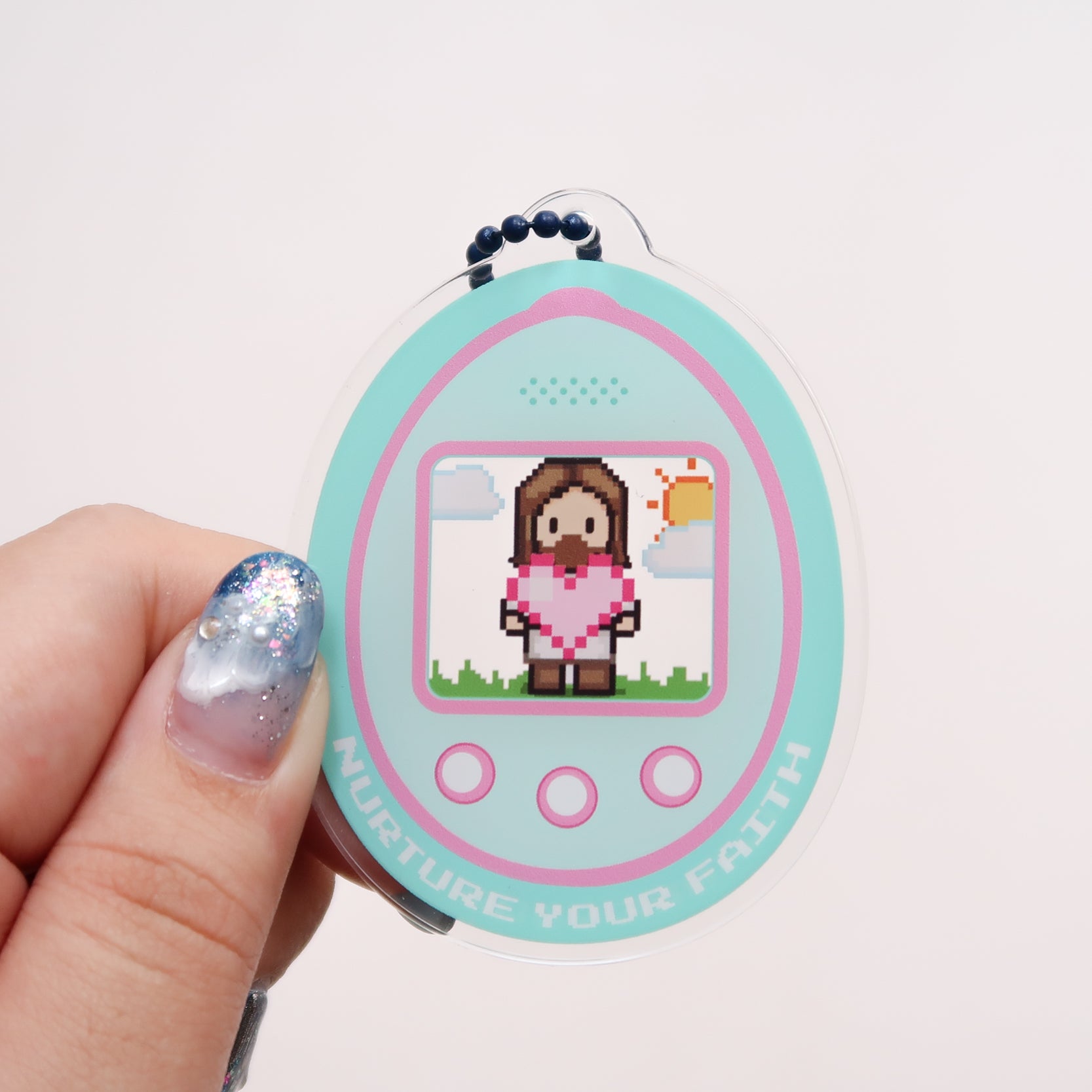 Oval acrylic keychain featuring a pixel-art digital pet with the message “NURTURE YOUR FAITH” at the bottom, attached to a blue ball chain