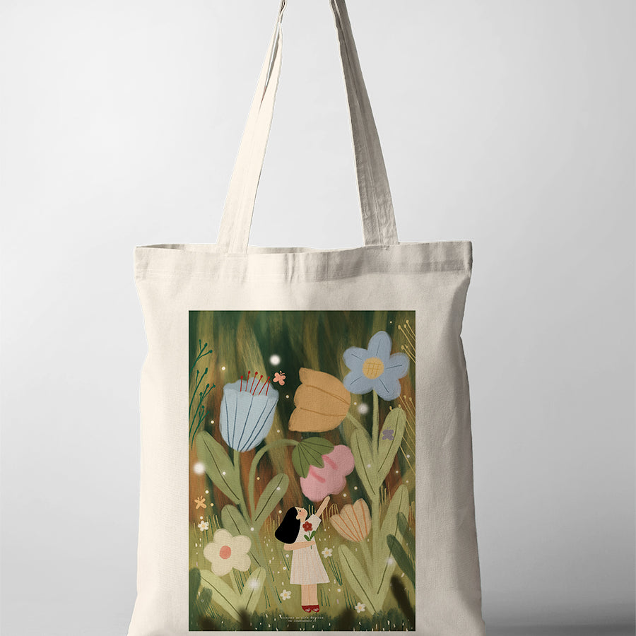 Canvas tote bag featuring Dita artwork of a girl standing among large whimsical flowers in a green field, soft storybook style illustration