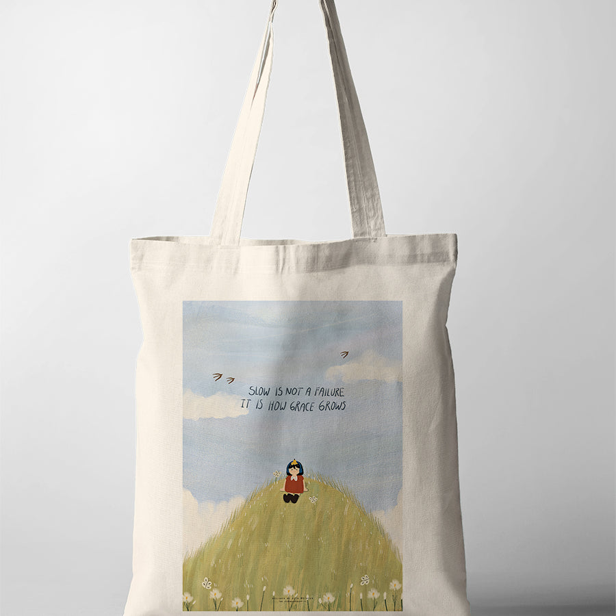 Canvas tote bag with Dita illustration of a girl sitting on a grassy hill with the phrase “Slow is not a failure, it is how grace grows” against a soft sky background