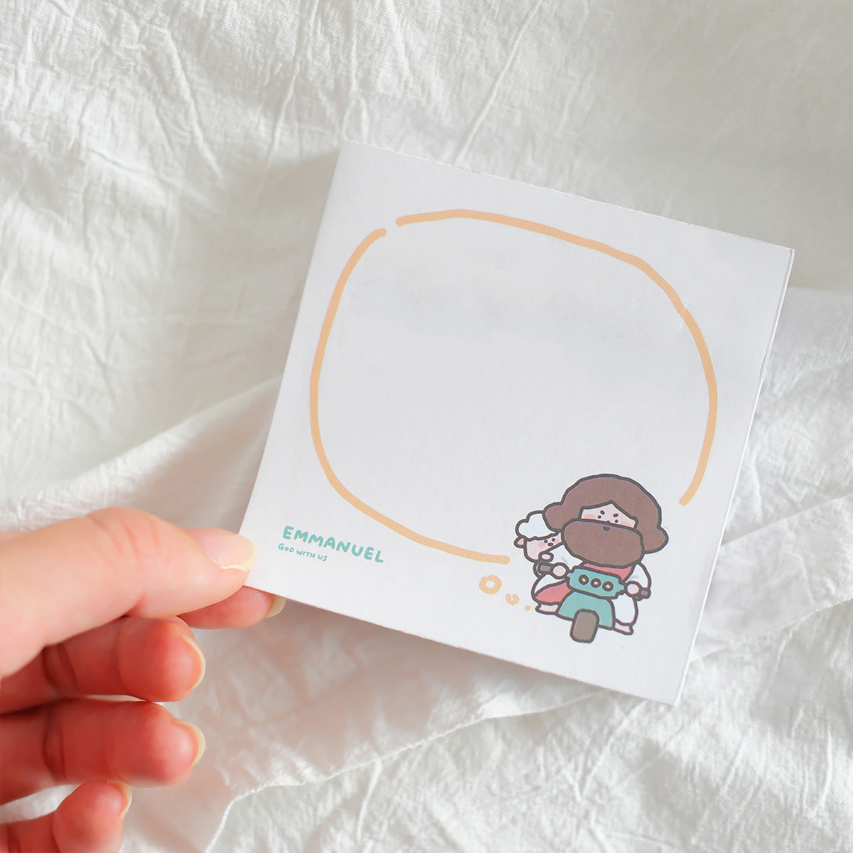 Hand holding a Christian-themed sticky note with a cute illustration of Jesus riding a scooter with a sheep, featuring the text 'Emmanuel, God with us' and a minimalist design for inspirational stationery.