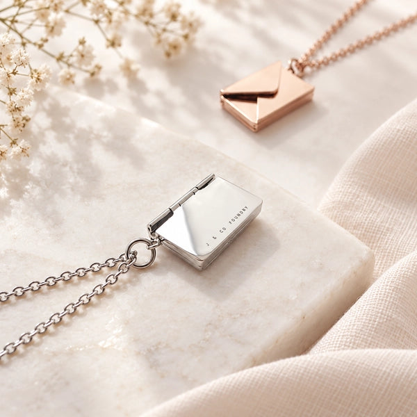 Silver and rose gold envelope locket necklaces styled on a light stone surface