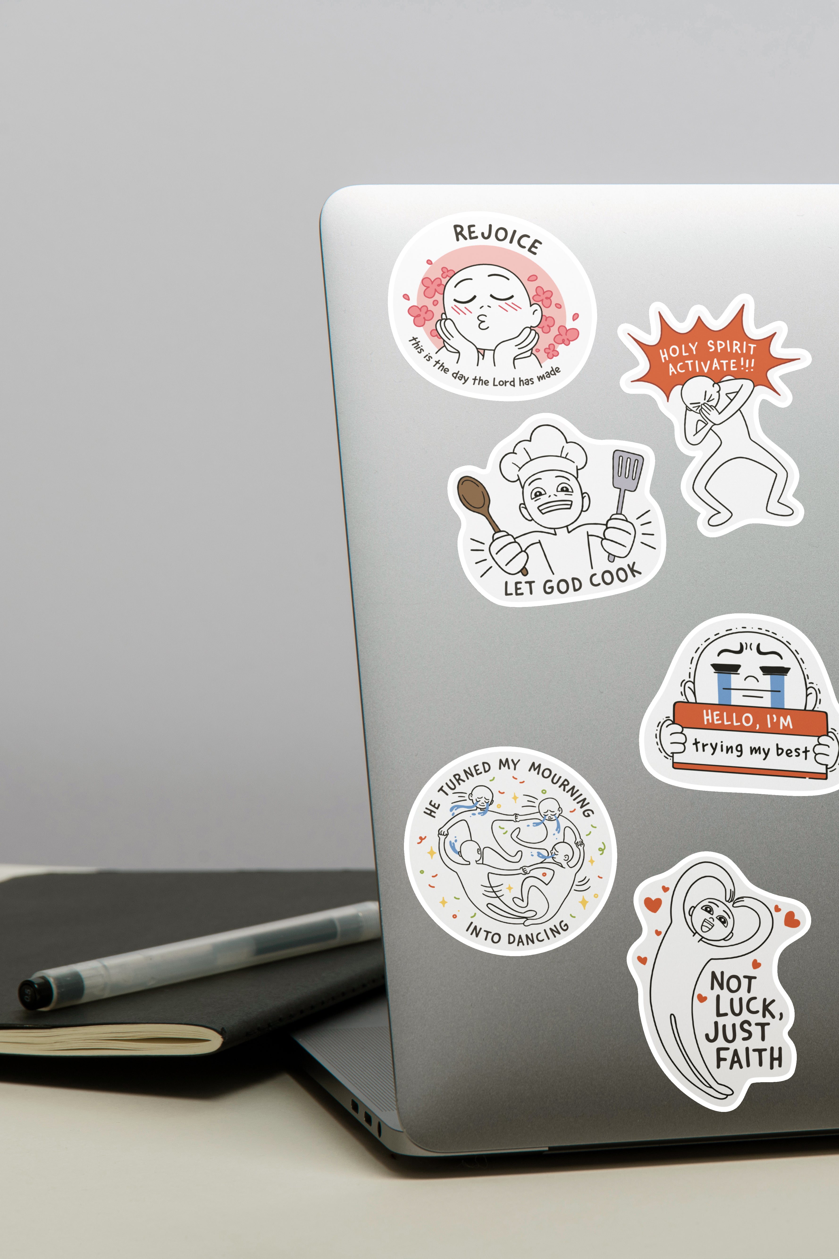 Laptop adorned with Everyday Believer stickers featuring Christian messages like "Rejoice," "Let God Cook," and "Not Luck, Just Faith," from The Commandment Co, Singapore.