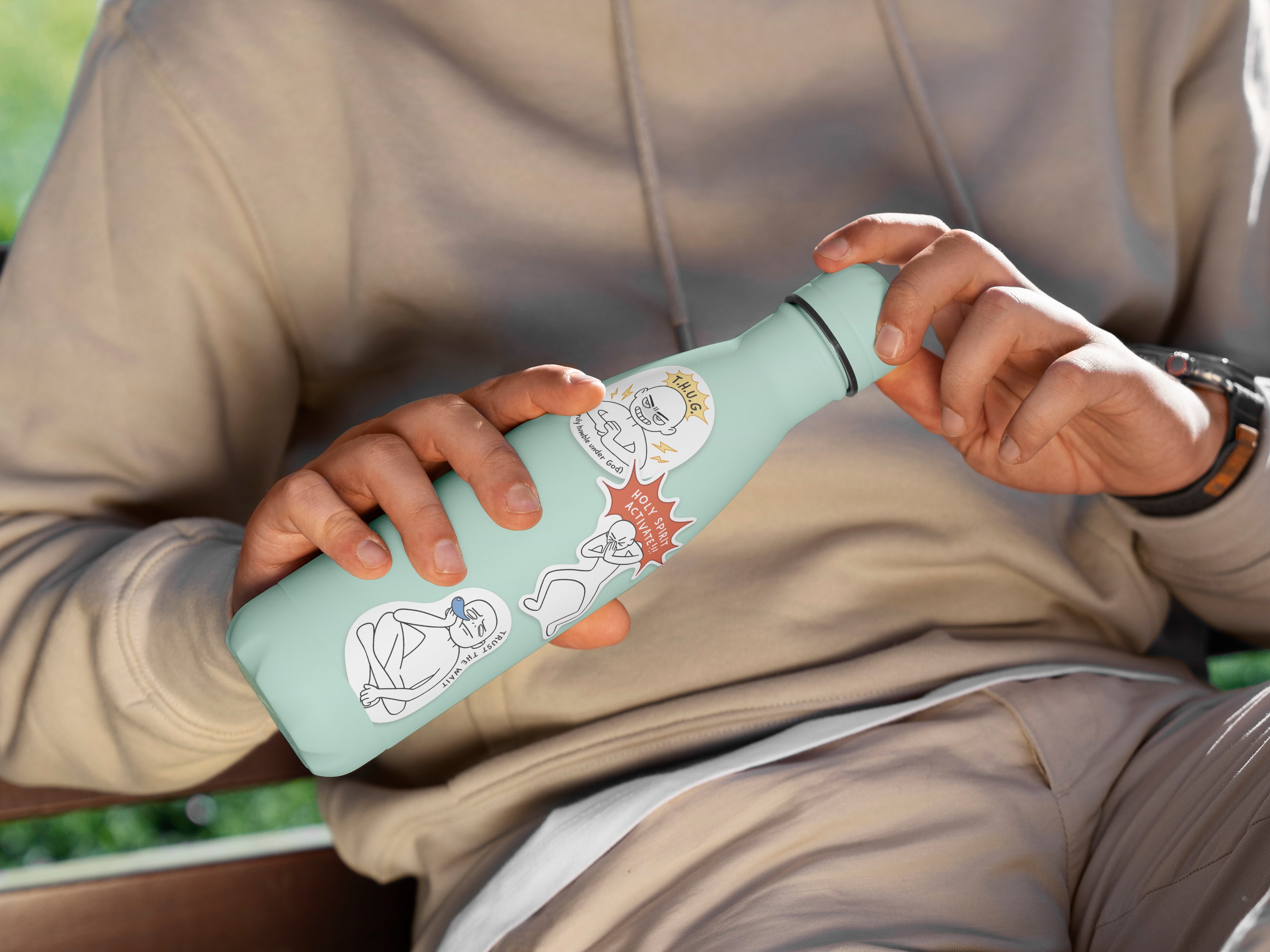 Person holding a water bottle decorated with Everyday Believer stickers featuring Christian messages like "Holy Spirit Activate" and "T.H.U.G.," from The Commandment Co, Singapore.