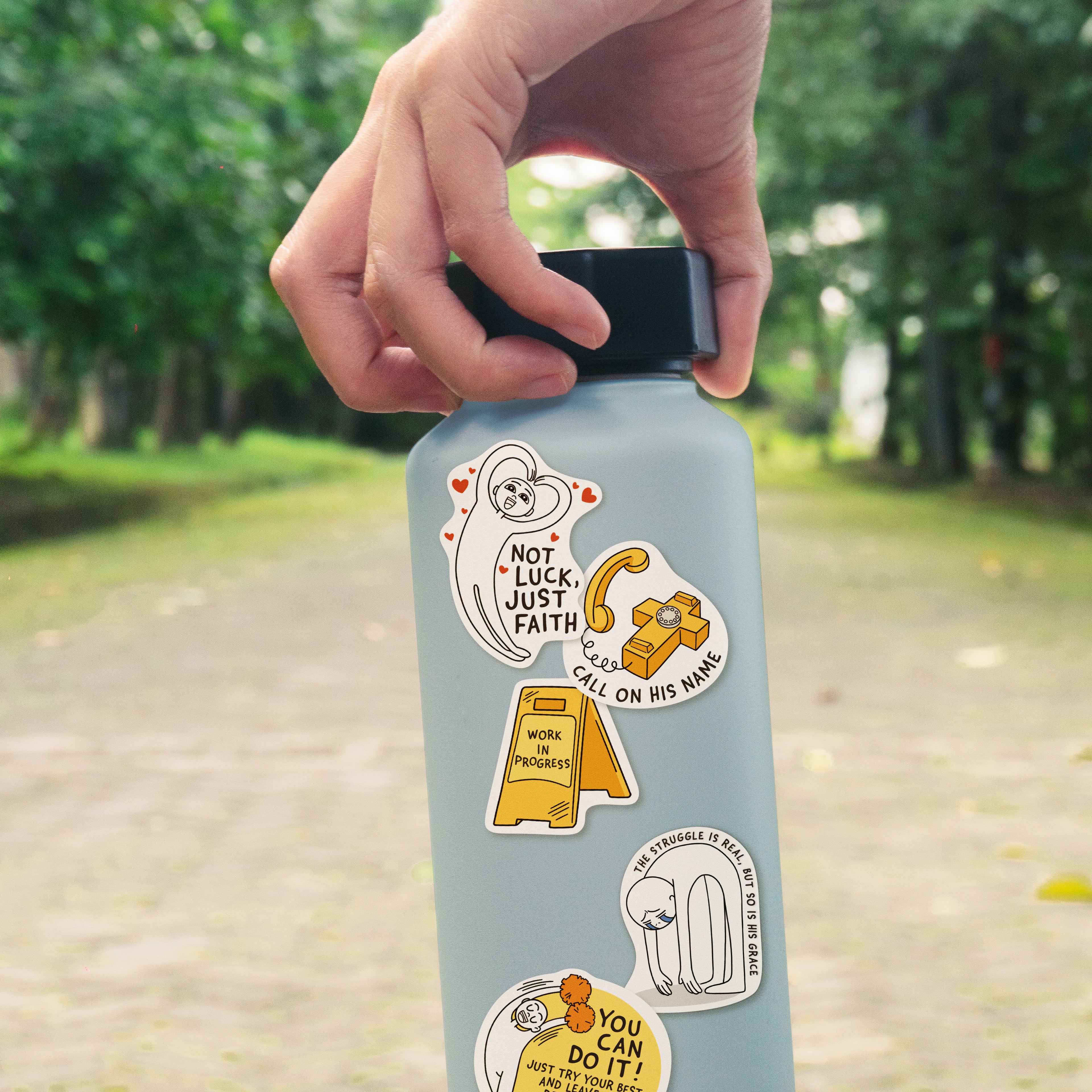 Hand holding a water bottle decorated with Everyday Believer stickers featuring Christian messages like "Not Luck, Just Faith" and "Call on His Name," from The Commandment Co, Singapore.